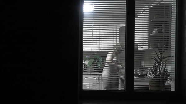 Peeping. Voyeur. Neighbor pervert voyeur in evening on street looks out window as naked sexy neighbor in kitchen prepares dinner for her husband. Naked in public. Naked at home. Family.  Outdoor