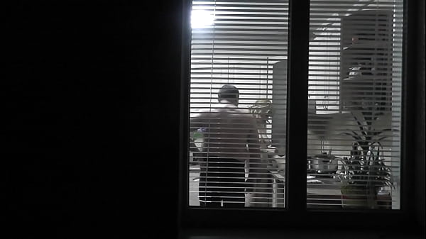 Peeping. Voyeur. Neighbor pervert voyeur in evening on street looks out window as naked sexy neighbor in kitchen prepares dinner for her husband. Naked in public. Naked at home. Family.  Outdoor
